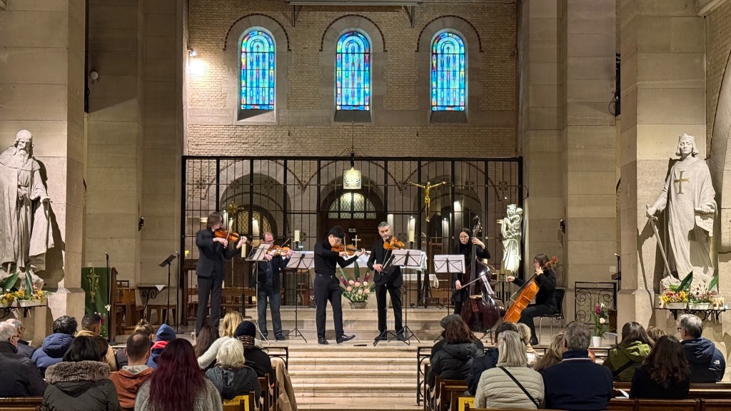 A string quartet performing in a church, with stained glass windows in the background and a statue on either side, while an audience listens attentively.